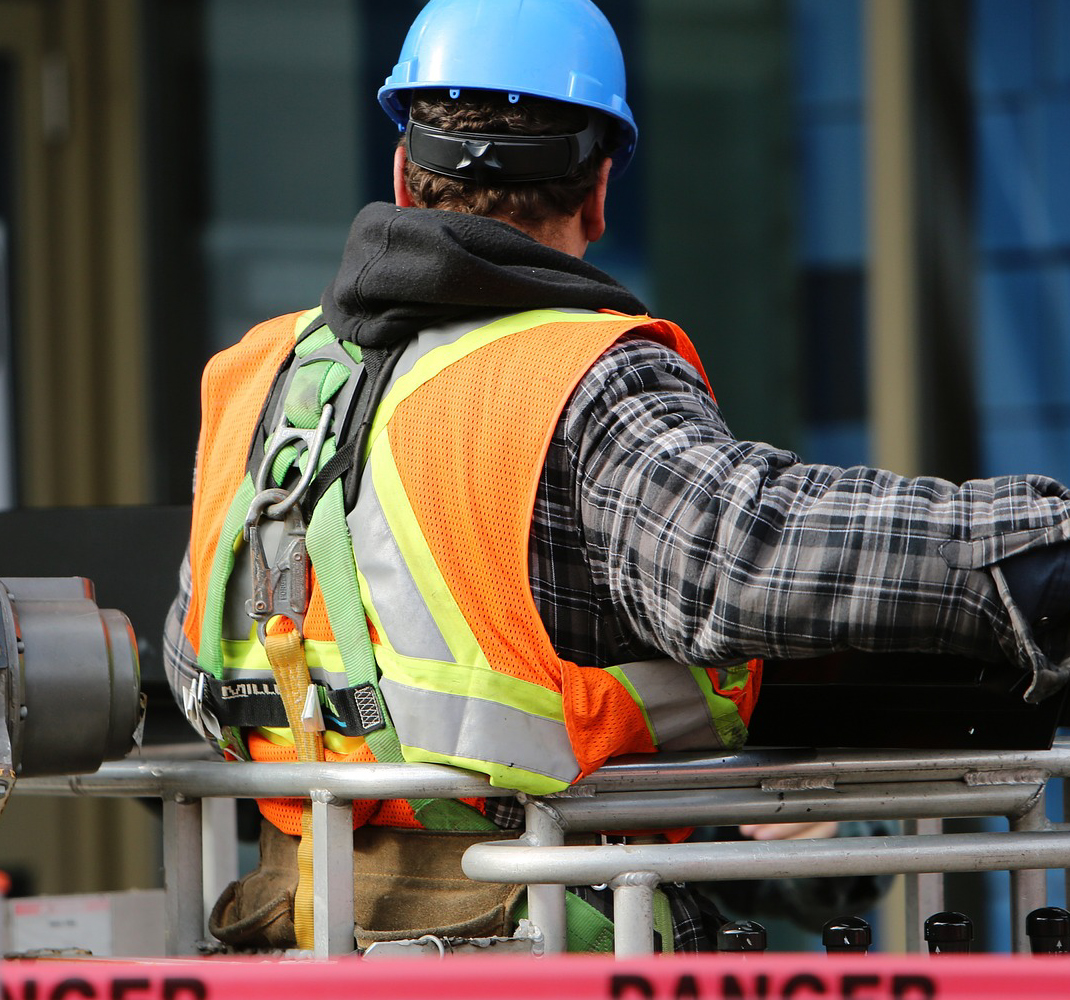 Image of a construction worker on a job site in a hi-vis orange vest wearing a hard hat on a lift